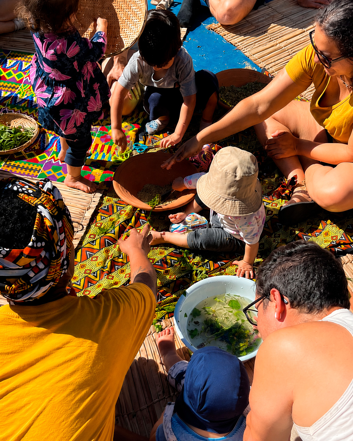 Vista aérea de adultos e crianças em roda sobre tecidos afro-brasileiros e esteiras, manipulando folhas e ervas verdes em bacias com água e barro em uma atividade sensorial ao ar livre.